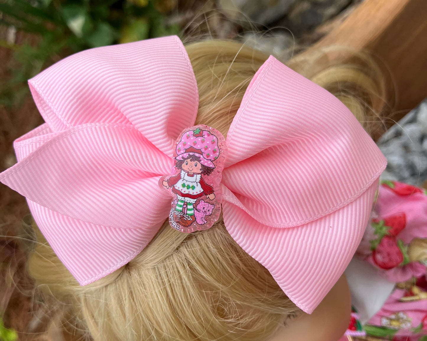 Close-up of a pink hair bow with a decorative clip on a person's head, with a blurred outdoor background.