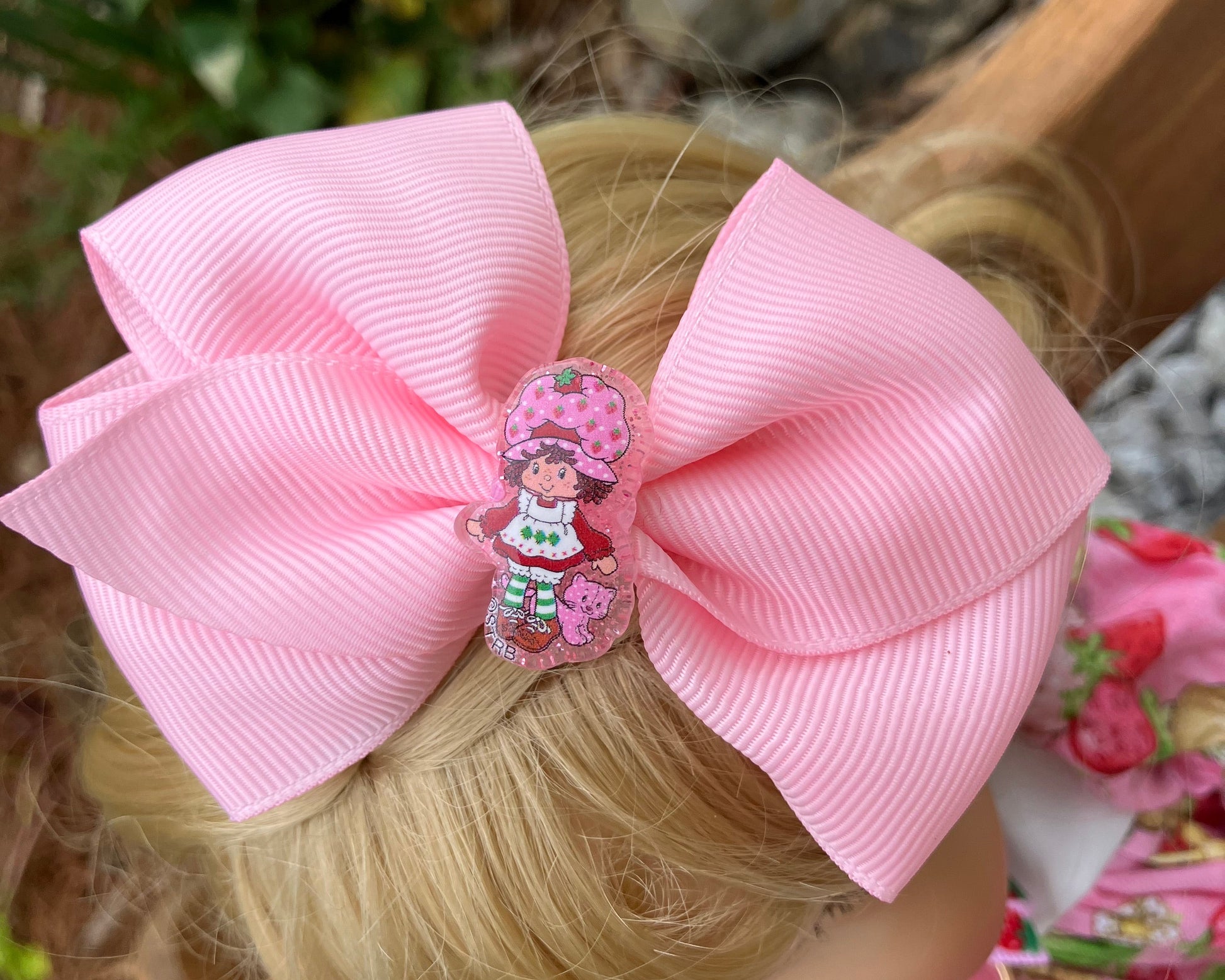Close-up of a pink hair bow with a decorative clip on a person's head, with a blurred outdoor background.